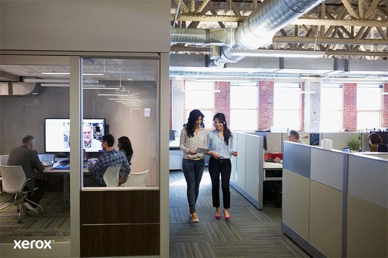 Deux femmes se promènent dans un espace de bureau ouvert moderne, engagées dans une conversation et entourées de postes de travail.