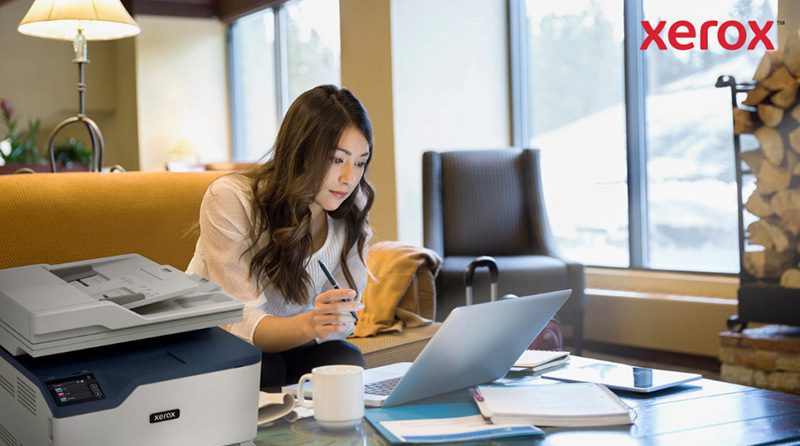 Plateforme Xerox® Workflow Central : travaillez n'importe où Une femme travaille sur un ordinateur portable dans un bureau ou un salon confortable, avec une imprimante Xerox compacte visible au premier plan, indiquant une intégration transparente de l'impression avec les flux de travail numériques.