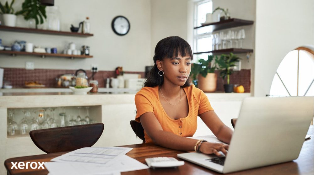 Xerox® DocuShare® Go : dynamiser le travail à distance Une personne est assise à une table de cuisine, concentrée sur son travail sur un ordinateur portable, avec des papiers et une calculatrice à proximité, illustrant la flexibilité du travail à domicile avec une gestion efficace des documents.