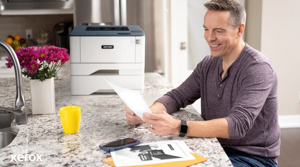 Expérience d'impression et de numérisation Xerox® : gestion simplifiée des documents Une personne sourit tout en examinant un document sur un comptoir de cuisine, avec une imprimante Xerox compacte et une tasse jaune vif à proximité, suggérant une impression et une gestion de documents transparentes dans un environnement domestique ou de petit bureau.